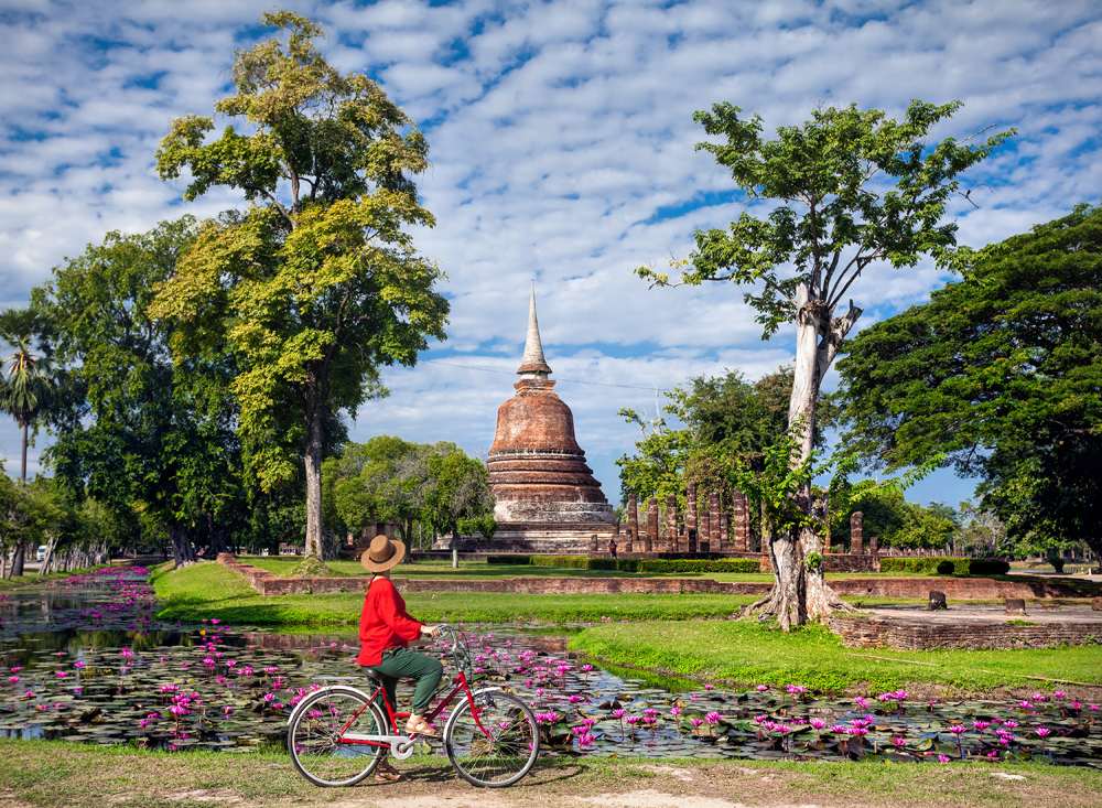 Woman riding bicycle and looking at old Buddhist temple in Sukhothai Historical Park, Thailand 