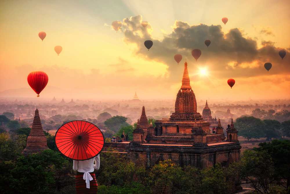 Burmese woman holding traditional red umbrella looking at Hot air balloon over Bagan in misty morning, Myanmar
